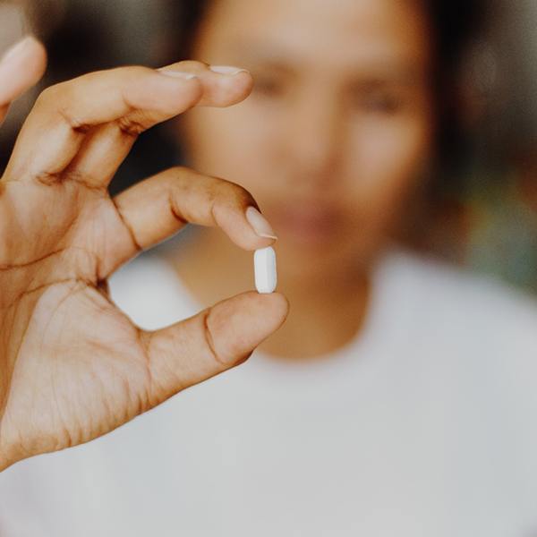 Woman blurry in background holding white pill between forefinger and thumb in foreground